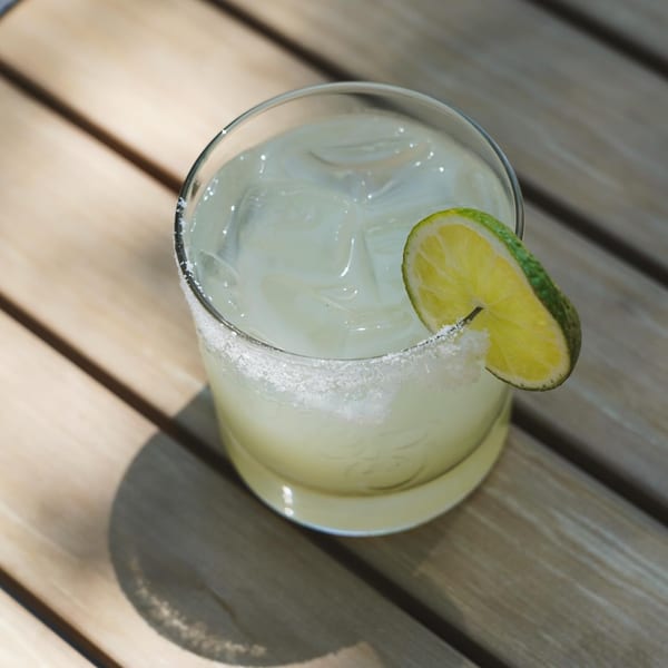 A margarita with a salted rim and lime wedge, on a wooden table in sunlight.