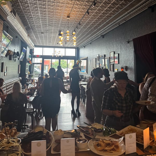 Guests mingle and dine in the long, dimly lit Luna & The Bear restaurant with a buffet table in the foreground.