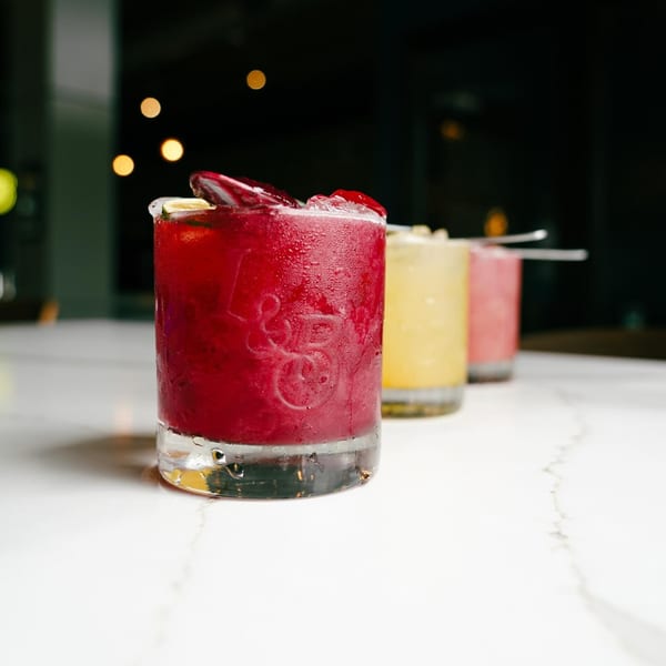 Three vibrant cocktails, red, yellow, and pink, with ice and garnishes on a white marble surface.