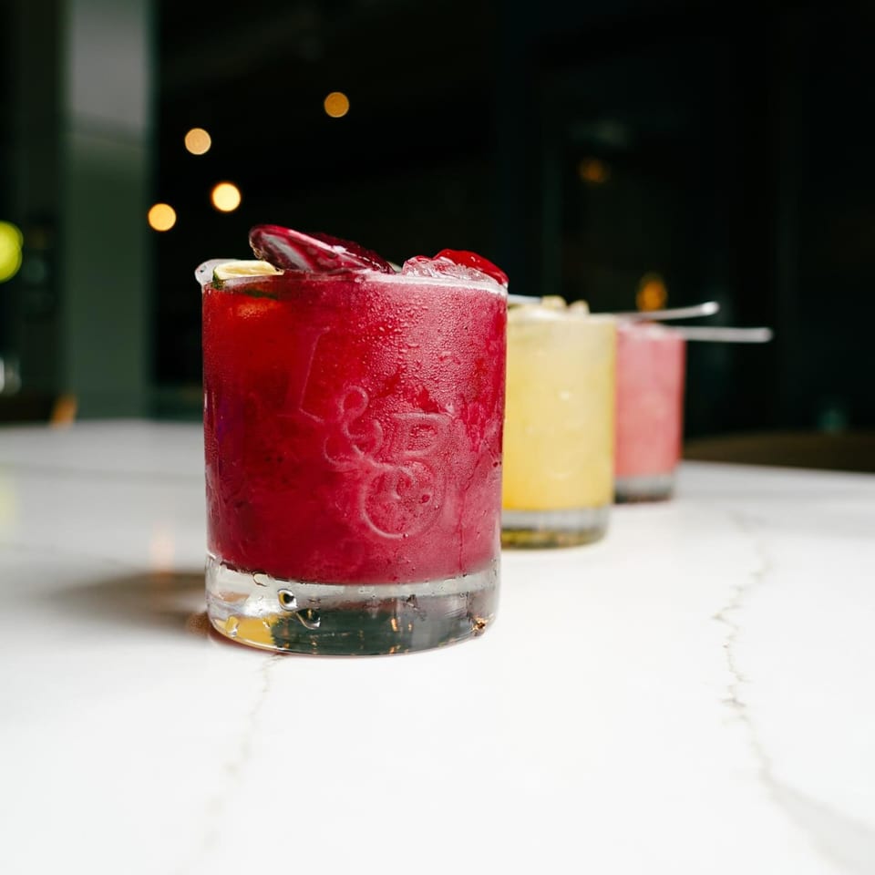 Three vibrant cocktails, red, yellow, and pink, with ice and garnishes on a white marble surface.