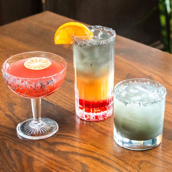 Three colorful cocktails, one pink, one layered orange and red, and one light blue, on a wooden table.