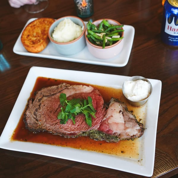 A prime rib entree with gravy, horseradish, and parsley, alongside sides of mashed potatoes, green beans, and a hash brown.