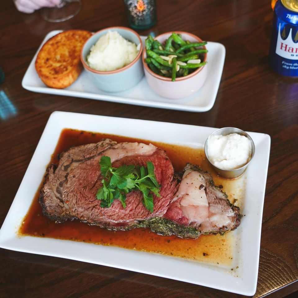 A prime rib entree with gravy, horseradish, and parsley, alongside sides of mashed potatoes, green beans, and a hash brown.