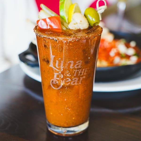 A Luna & The Bear Bloody Mary cocktail, garnished with lime, olives, and peppers, on a dark table.