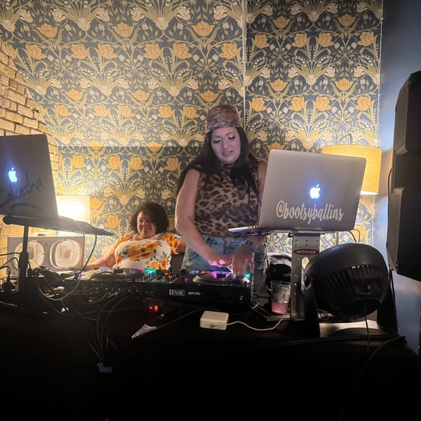 Two women DJing at Luna & The Bear, with laptops and turntables against floral wallpaper.
